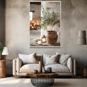 May include: A rustic wooden vase with a green olive branch inside, sitting on a table with two lit candles and a lantern. The vase is in front of a white wall with a window in the background.