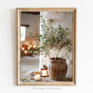 May include: A rustic wooden vase with a green olive branch inside, sitting on a table with two lit candles and a wooden lantern. The vase is in a whitewashed room with a wooden frame around the image.
