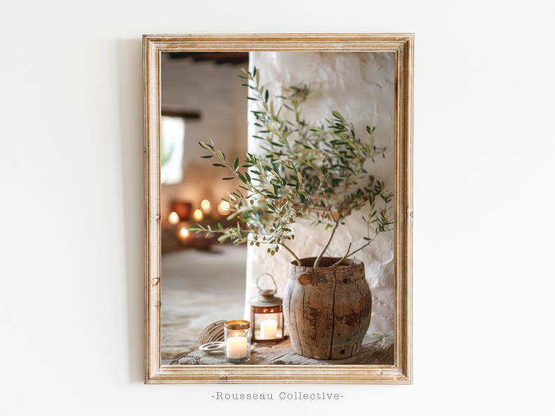 May include: A rustic wooden vase with a green olive branch inside, sitting on a table with two lit candles and a wooden lantern. The vase is in a whitewashed room with a wooden frame around the image.