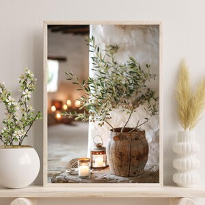 May include: A rustic wooden vase with a green olive branch plant inside, sitting on a wooden table with two candles and a lantern. The vase is surrounded by a white wall and a window with a blurry view of a room.
