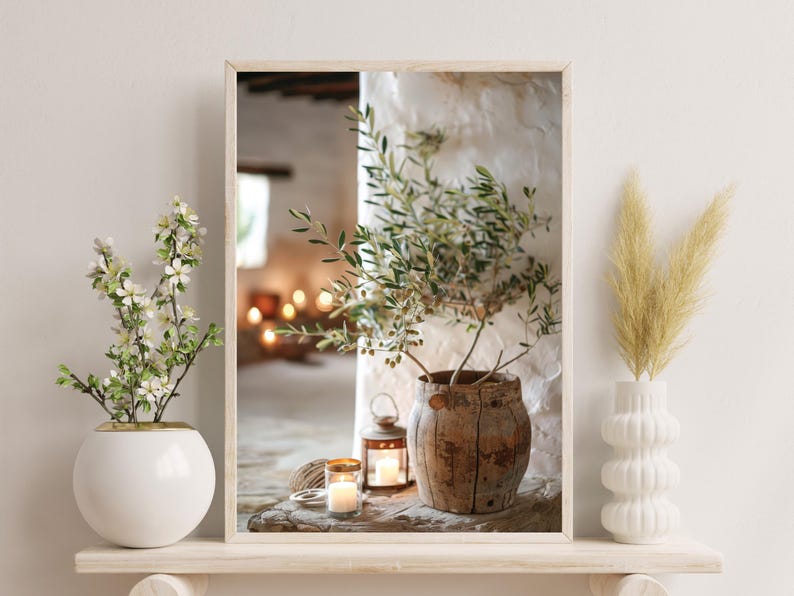 May include: A rustic wooden vase with a green olive branch plant inside, sitting on a wooden table with two candles and a lantern. The vase is surrounded by a white wall and a window with a blurry view of a room.