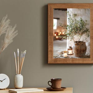 May include: A framed photograph of a rustic interior with a potted olive branch, candles, and a lantern. The photo is in a wooden frame with a light brown finish.