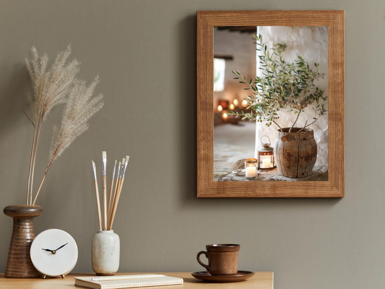 May include: A framed photograph of a rustic interior with a potted olive branch, candles, and a lantern. The photo is in a wooden frame with a light brown finish.