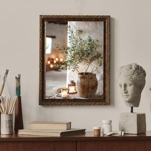 May include: A framed photograph of a rustic still life with a potted olive branch, a lantern, and candles. The photograph is displayed on a wooden shelf with other decorative items, including a bust of a woman and a wooden hand.
