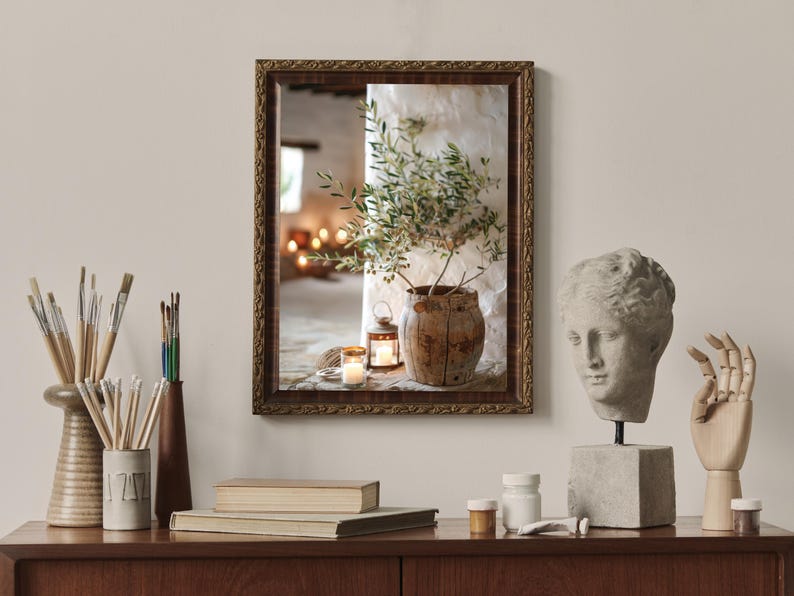 May include: A framed photograph of a rustic still life with a potted olive branch, a lantern, and candles. The photograph is displayed on a wooden shelf with other decorative items, including a bust of a woman and a wooden hand.