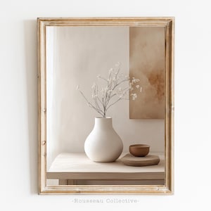 Könnte beinhalten: Eine weiße Keramikvase mit einem zarten Zweig weißer Blumen steht auf einem Holztisch vor einem beige-braunen abstrakten Gemälde. Die Vase und die Blumen spiegeln sich in einem großen, gerahmten Spiegel.