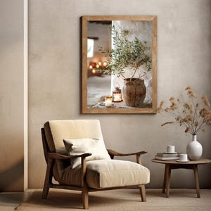May include: A framed photograph of a potted olive branch with candles in a rustic setting. The photograph is hanging on a wall in a room with a beige armchair and a side table with a vase of dried flowers.