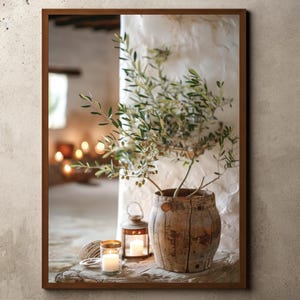 May include: A rustic wooden pot with a small olive tree inside, sitting on a wooden surface with two candles and a lantern. The background is a white wall with a window.