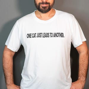 以下が含まれることがあります： 黒い文字で「ONE CAT JUST LEADS TO ANOTHER.」とプリントされた白いTシャツです。
