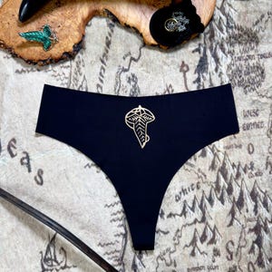 以下が含まれることがあります： 金色の葉のデザインが施された黒い下着。下着は中つ国の地図の上に置かれています。