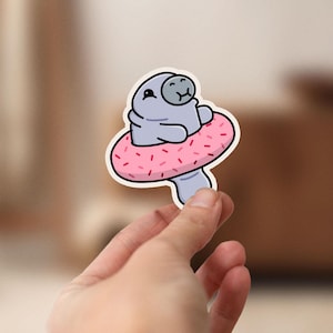 May include: A sticker of a cartoon manatee wearing a pink donut-shaped pool float. The manatee is gray and has a happy expression. The donut has pink frosting and red sprinkles.