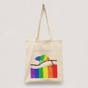 Op de afbeelding: Een beige tote bag met een regenboogvlag en een kameleon. De tekst "Verberg je niet, toon je trots!" staat op de tas gedrukt.