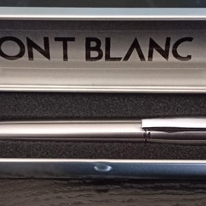 Może przedstawiać: Srebrny długopis Montblanc w srebrnym etui. Pióro spoczywa na czarnej piance wewnątrz etui. Logo i nazwa marki Montblanc są wydrukowane na czarno wewnątrz etui.