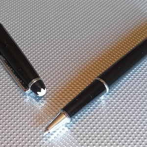 Può includere: Una penna roller nera con punta e dettagli argentati, e un cappuccio staccato. Il cappuccio presenta una fascia argentata e un'emblema bianco. La penna è su una superficie argentata strutturata. È uno strumento di scrittura.