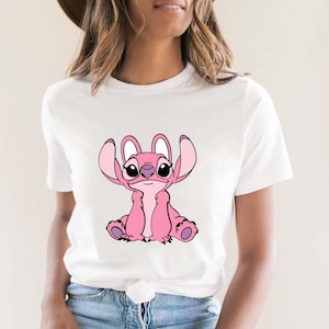 May include: White t-shirt featuring a pink cartoon character with large ears and a friendly expression. The character is sitting, with its arms and legs visible. The design is centered on the shirt.