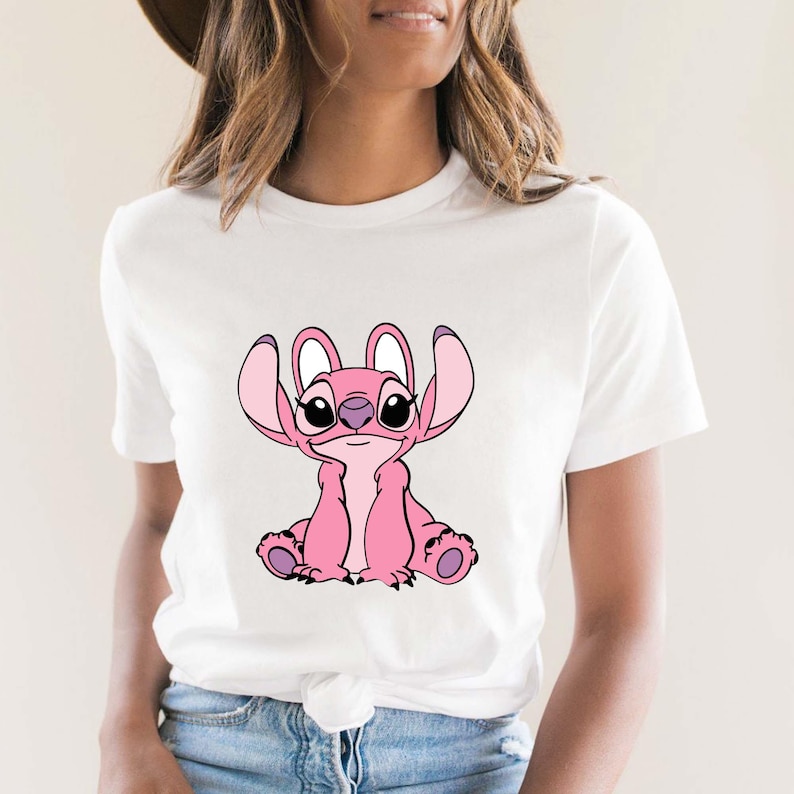 May include: White t-shirt featuring a pink cartoon character with large ears and a friendly expression. The character is sitting, with its arms and legs visible. The design is centered on the shirt.