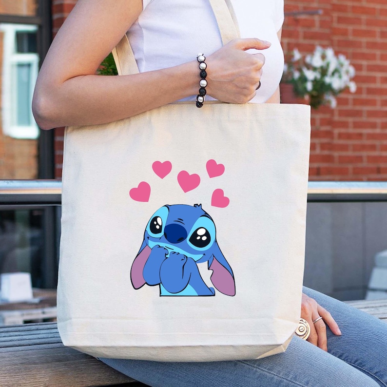 May include: Cream-colored tote bag featuring a blue cartoon character with large eyes and pink ears, surrounded by pink hearts. The bag has a simple design with a rectangular shape and long straps.