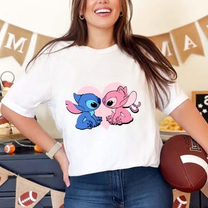 May include: White t-shirt featuring a cartoon graphic of two characters, one blue and one pink, facing each other in front of a pink heart. The blue character has large ears and the pink character has long, curly ears. The shirt has short sleeves.