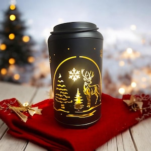 May include: A black metal lantern with a lid, featuring a cut-out design of a deer, trees, and a snowflake, illuminated with a warm glow. The lantern sits on a red cloth, with a blurred background of a Christmas tree and lights.