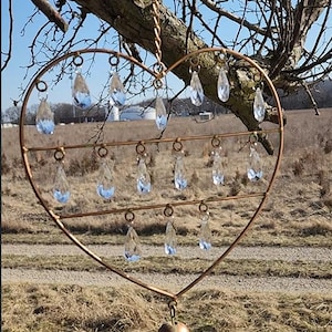 May include: A heart-shaped copper wind chime with clear, teardrop-shaped crystal pendants. A small bell hangs at the bottom, suspended by a chain. The chime is hanging from a tree branch against a backdrop of a field and blue sky.