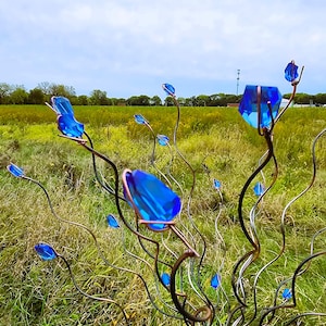 May include: A metal sculpture featuring blue glass crystal accents, resembling stylized flowers or buds. The sculpture has a dark metal frame with curved branches, set against a backdrop of a grassy field and a cloudy sky.