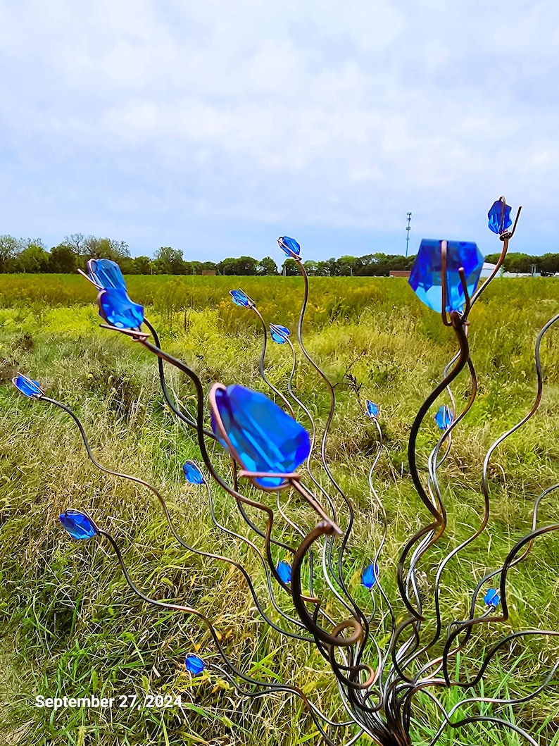 May include: A metal sculpture featuring blue glass crystal accents, resembling stylized flowers or buds. The sculpture has a dark metal frame with curved branches, set against a backdrop of a grassy field and a cloudy sky.