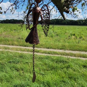 Pu&ograve; includere: Un carillon a vento in metallo color rame a forma di angelo con dettagli intricati e una catena che pende dal fondo. Il carillon a vento &egrave; appeso a un ramo d'albero.