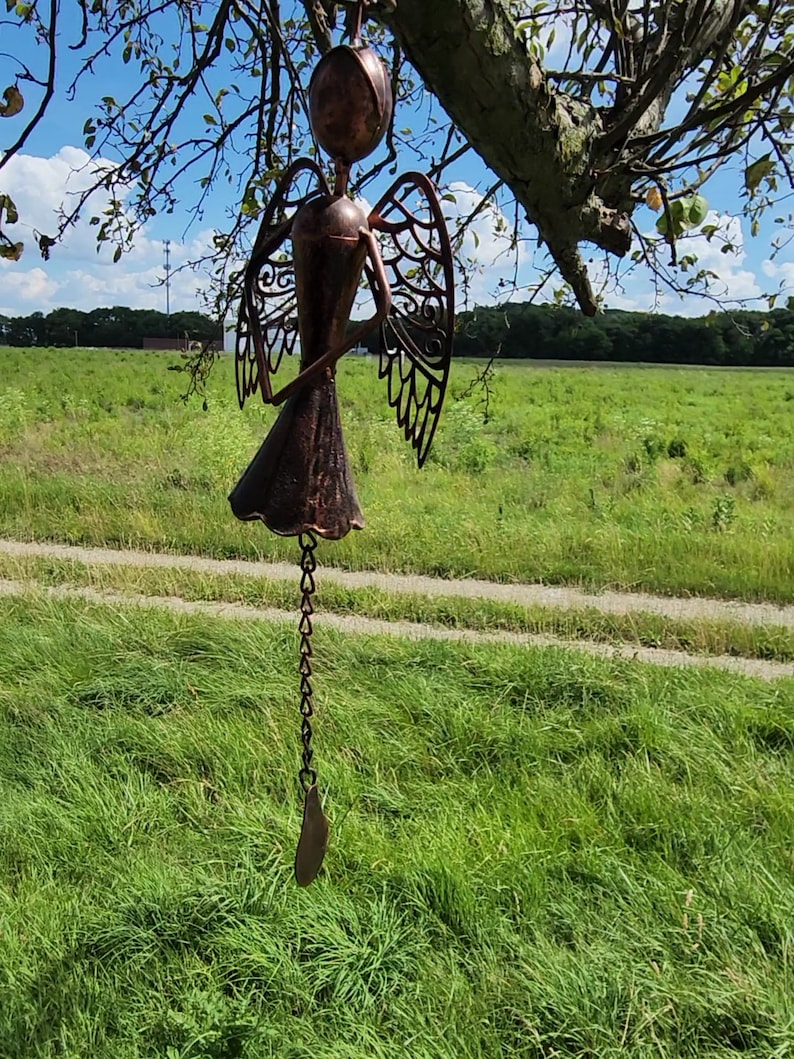 Pu&ograve; includere: Un carillon a vento in metallo color rame a forma di angelo con dettagli intricati e una catena che pende dal fondo. Il carillon a vento &egrave; appeso a un ramo d'albero.