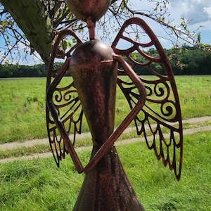 Pu&ograve; includere: Una statuetta di angelo in metallo color rame con ali tagliate. L'angelo &egrave; in piedi in un campo erboso con un albero sullo sfondo.