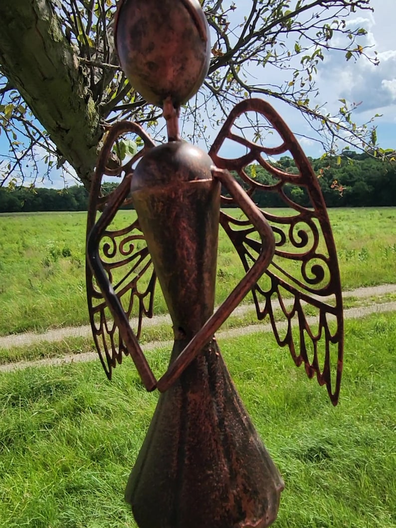 Pu&ograve; includere: Una statuetta di angelo in metallo color rame con ali tagliate. L'angelo &egrave; in piedi in un campo erboso con un albero sullo sfondo.