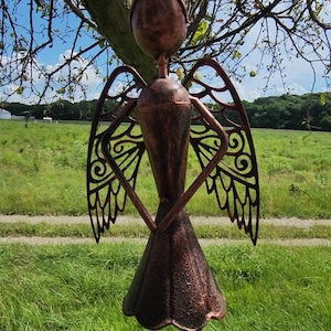 Pu&ograve; includere: Un carillon a vento in metallo color rame a forma di angelo con ali tagliate e una campana in basso. L'angelo &egrave; appeso a un ramo d'albero.