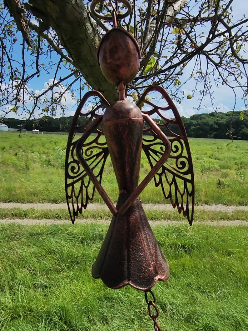 Pu&ograve; includere: Un carillon a vento in metallo color rame a forma di angelo con ali tagliate e una campana in basso. L'angelo &egrave; appeso a un ramo d'albero.