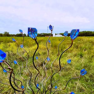 May include: A metal sculpture of stylized flowers with blue glass petals. The stems are dark and winding, with the blue glass pieces at the tips. The sculpture is set in a field of green and yellow grass under a cloudy sky.