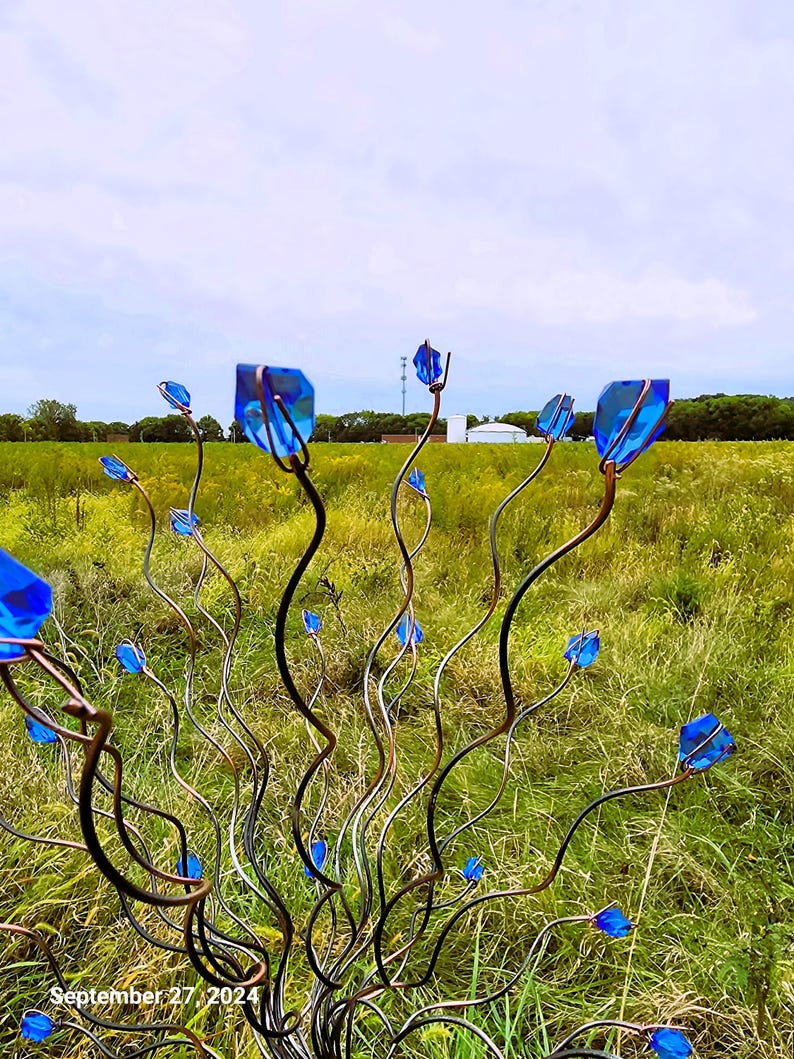 May include: A metal sculpture of stylized flowers with blue glass petals. The stems are dark and winding, with the blue glass pieces at the tips. The sculpture is set in a field of green and yellow grass under a cloudy sky.