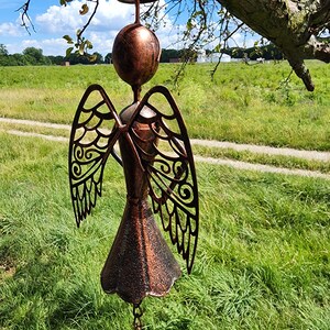 Pu&ograve; includere: Un carillon a vento in metallo color rame a forma di angelo con ali tagliate intricate e una campana nella parte inferiore. L'angelo &egrave; appeso a una catena ed &egrave; sospeso di fronte a un campo erboso verde.