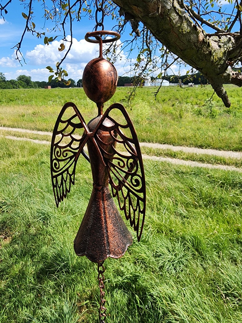 Pu&ograve; includere: Un carillon a vento in metallo color rame a forma di angelo con ali tagliate intricate e una campana nella parte inferiore. L'angelo &egrave; appeso a una catena ed &egrave; sospeso di fronte a un campo erboso verde.