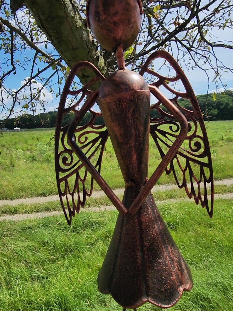 Pu&ograve; includere: Una scultura di angelo in metallo con ali intricate e una finitura in rame anticato. L'angelo &egrave; in piedi in un campo erboso.