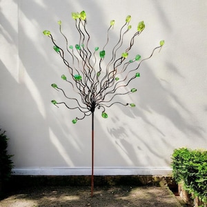 May include: A decorative metal tree sculpture with a brown stem and dark, wavy branches. The branches are adorned with green, gem-like accents. The sculpture stands in a garden setting against a white wall.