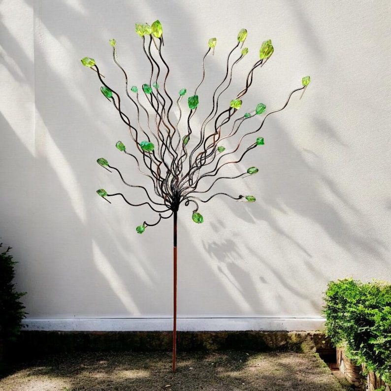 May include: A decorative metal tree sculpture with a brown stem and dark, wavy branches. The branches are adorned with green, gem-like accents. The sculpture stands in a garden setting against a white wall.