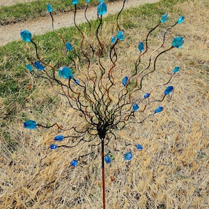 May include: A decorative garden stake featuring a central stem with a network of dark brown, twisting branches. Each branch holds a vibrant blue glass flower. The stake is set in a field of dry grass.