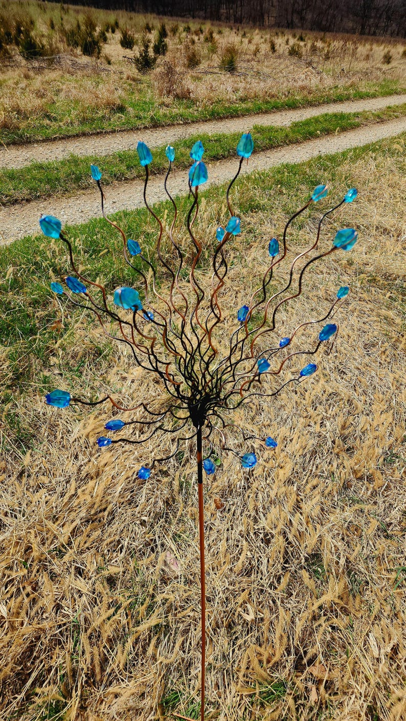 May include: A decorative garden stake featuring a central stem with a network of dark brown, twisting branches. Each branch holds a vibrant blue glass flower. The stake is set in a field of dry grass.