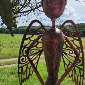 Pu&ograve; includere: Una scultura da giardino in metallo di un angelo con le ali. L'angelo &egrave; fatto di metallo marrone arrugginito e ha un design dettagliato. La scultura &egrave; in piedi in un campo erboso.