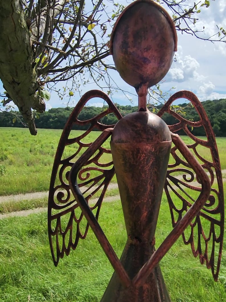 Pu&ograve; includere: Una scultura da giardino in metallo di un angelo con le ali. L'angelo &egrave; fatto di metallo marrone arrugginito e ha un design dettagliato. La scultura &egrave; in piedi in un campo erboso.