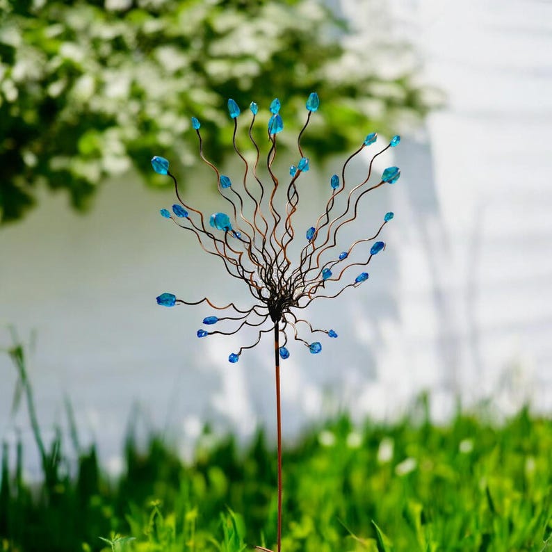 May include: A decorative metal garden stake featuring a cluster of wavy copper wires with blue glass bead accents. The stake has a long, slender stem and is designed to be placed in a garden or outdoor setting. The background is blurred, with green grass and foliage.