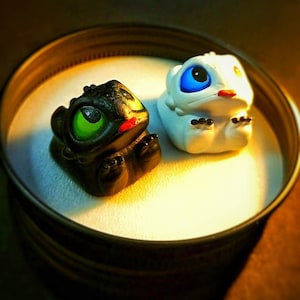 Puede incluir: Dos pequeñas figuritas de dragones pintadas a mano. Una es negra con ojos verdes y boca roja, la otra es blanca con ojos azules y boca roja. Están sentadas sobre una superficie blanca.