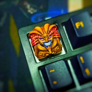 May include: A custom keycap with a detailed design of a character in shades of gold, red, and blue, set on a black keyboard. The F1 key is visible in the background.