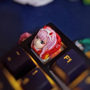 May include: A pink and red anime character keycap with a white headband and blue eyes. The keycap is on a black keyboard with a yellow accent.