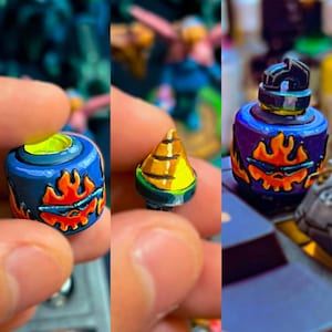 Pode incluir: Um pequeno componente cilíndrico azul para miniaturas de jogos com um design pintado de uma caveira em chamas laranja com óculos de sol. A peça tem um interior amarelo brilhante e é mostrada ao lado de um topo cónico amarelo e uma versão montada.