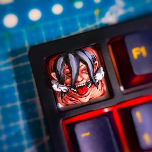 Pode incluir: Uma tecla artesanal personalizada para teclado mecânico com um personagem esculpido e pintado à mão, apresentando cabelos escuros, olhos vermelhos e boca aberta com a língua visível. A tecla está inserida num teclado preto ao lado da tecla F1.