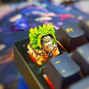 Puede incluir: Un tapón de tecla de anime verde y amarillo con una expresión feroz. El tapón de tecla está en un teclado negro.